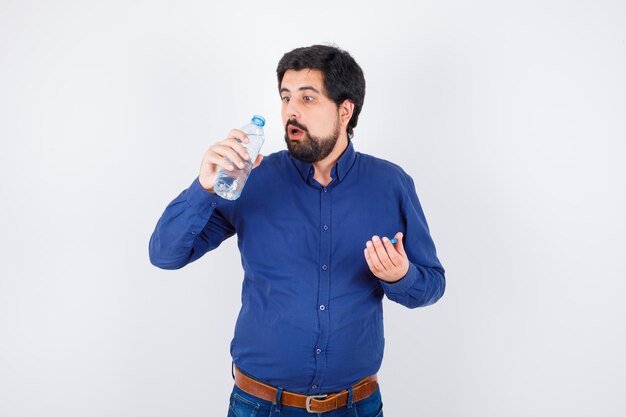 Jonge man die een waterfles opent en water probeert te drinken in een blauw shirt en spijkerbroek en er verrast uitziet, vooraanzicht.