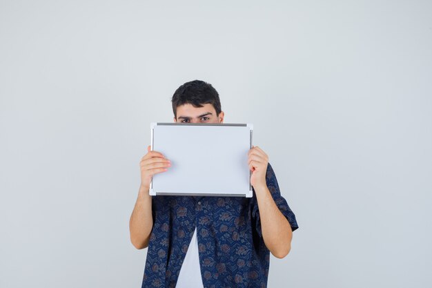 Jonge man die een whiteboard vasthoudt, zijn gezicht erachter verbergt in een wit t-shirt en een gebloemd hemd