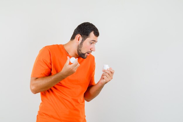 Jonge man die fles pillen in oranje t-shirt onderzoekt en gericht, vooraanzicht kijkt.