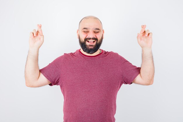 Jonge man die gekruiste vingers opsteekt terwijl hij lacht in een roze t-shirt en er vrolijk uitziet. vooraanzicht.