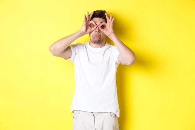 Jonge man die grappige gezichten maakt en tong laat zien, gek rond, staande in wit t-shirt tegen gele achtergrond