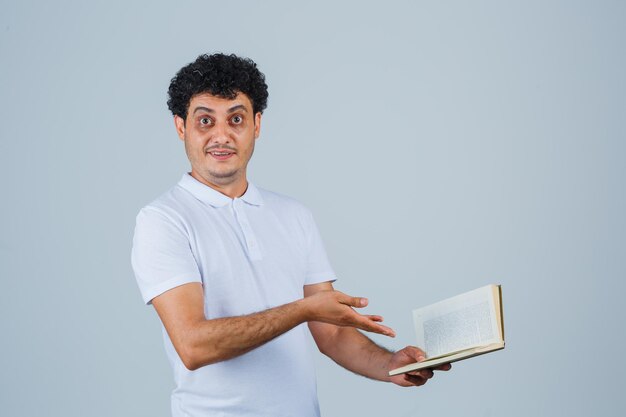 Jonge man die hand uitrekt naar boek in wit t-shirt en spijkerbroek en er gelukkig uitziet. vooraanzicht.