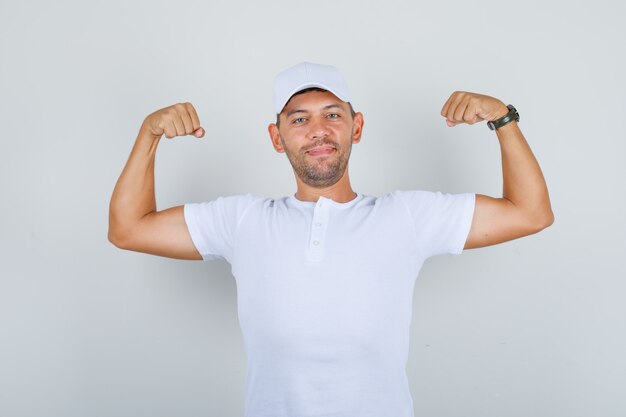 Jonge man die handen opheft en spieren in wit t-shirt, pet toont en sterk, vooraanzicht kijkt
