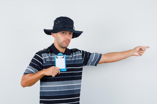 Jonge man die klembord vasthoudt terwijl hij naar de zijkant wijst in een t-shirt, hoed vooraanzicht.