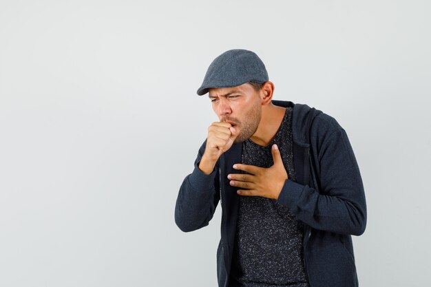 Jonge man die lijden aan hoest in t-shirt, jasje, pet en onwel kijkt