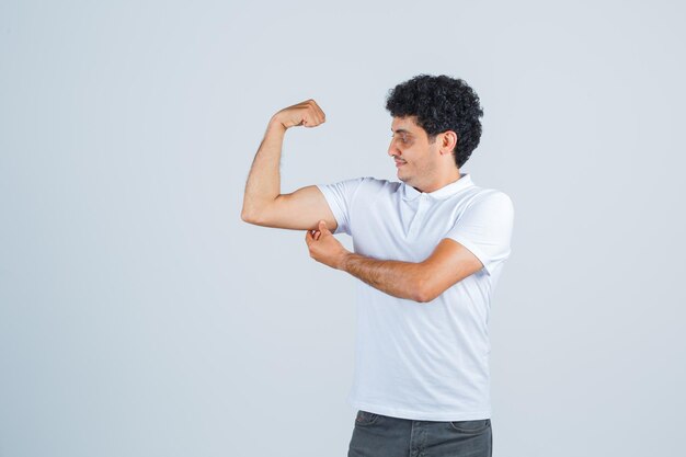 Jonge man die machtsgebaar toont, ernaar kijkt in een wit t-shirt en spijkerbroek en er gelukkig uitziet. vooraanzicht.
