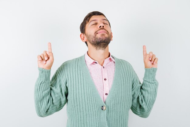 Jonge man die naar boven wijst, ogen dicht houdt in shirt, vest en dankbaar kijkt. vooraanzicht.