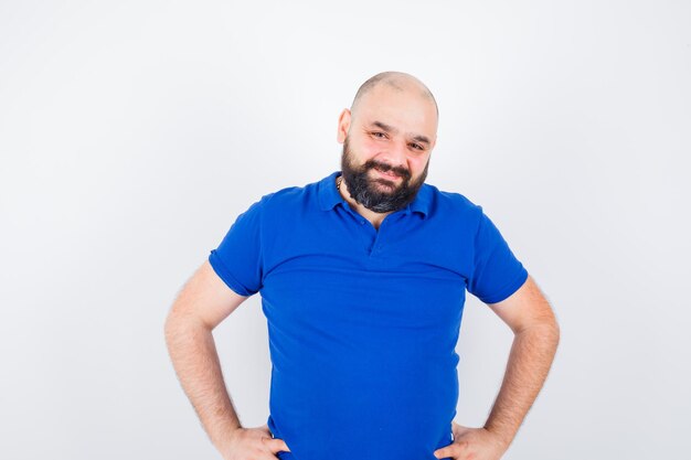 Jonge man die naar de camera kijkt met de handen op de taille terwijl hij lacht in een blauw shirt en er blij uitziet. vooraanzicht.