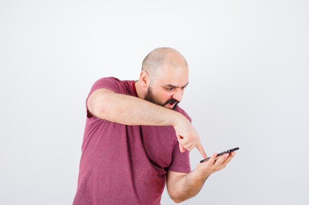 Jonge man die naar de telefoon kijkt en de wijsvinger ernaar wijst in een roze t-shirt en er boos uitziet. vooraanzicht.