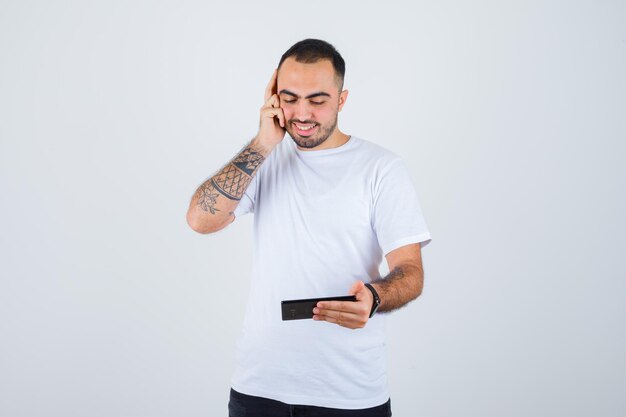 Jonge man die naar iets op de telefoon kijkt en de hand op het oor drukt in een wit t-shirt en een zwarte broek en er gelukkig uitziet