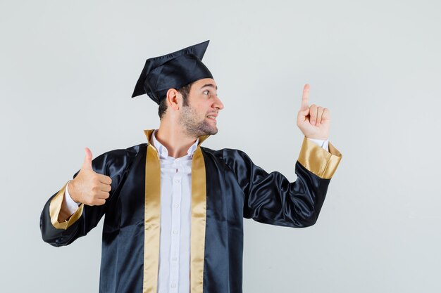 Jonge man die omhoog wijst, duim in gediplomeerde uniform toont en hoopvol kijkt. vooraanzicht.