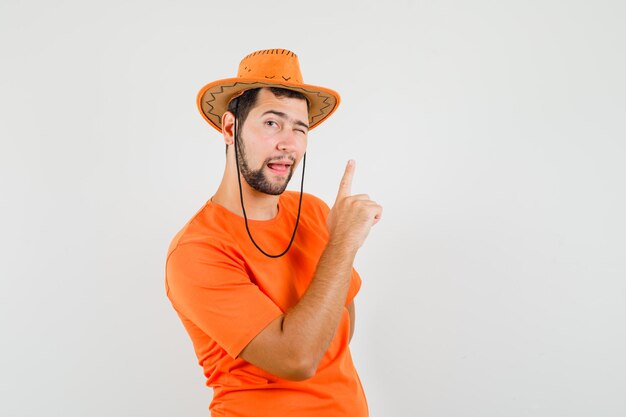 Jonge man die omhoog wijst, knipogend oog in oranje t-shirt, hoed, vooraanzicht.