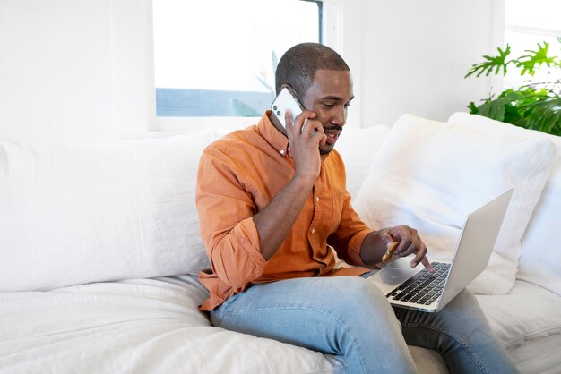Jonge man die op smartphone praat terwijl hij thuis een laptop gebruikt