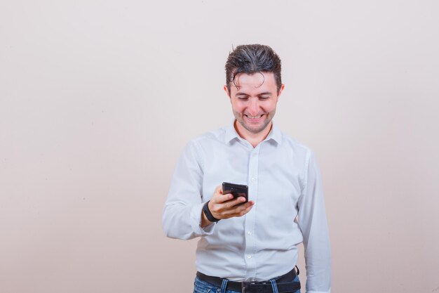 Jonge man die smartphone in shirt, spijkerbroek gebruikt en er vrolijk uitziet