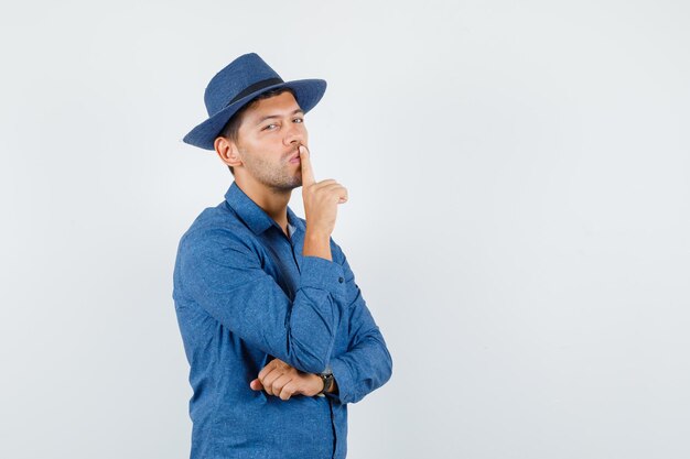 Jonge man die stiltegebaar in blauw shirt, hoed toont en er voorzichtig uitziet. vooraanzicht.