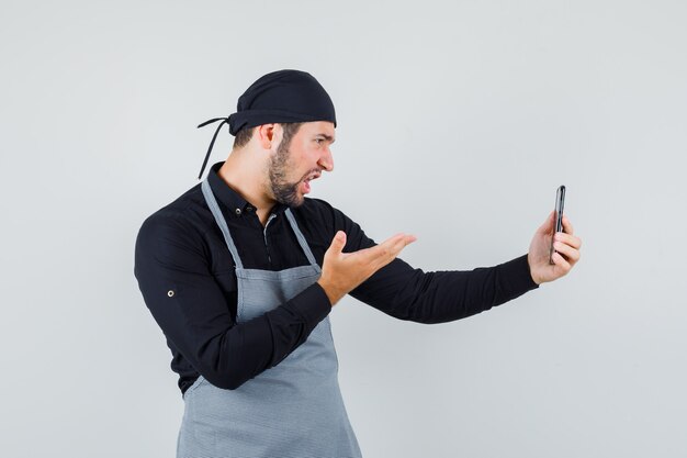 Jonge man die videogesprek voert op mobiele telefoon in overhemd, schort en woedend, vooraanzicht kijkt.