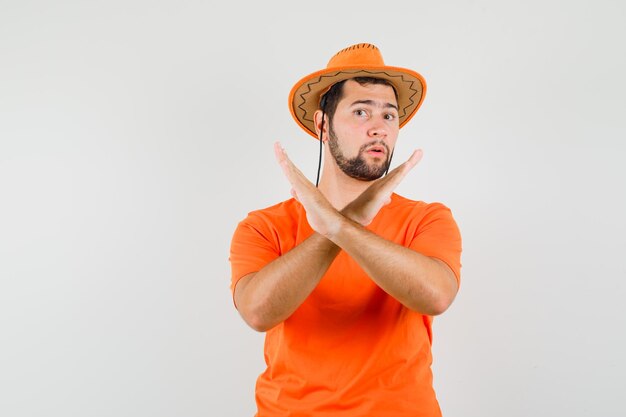 Jonge man die weigeringsgebaar toont in oranje t-shirt, hoed en er beslissend uitziet, vooraanzicht.