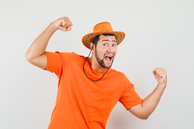 Jonge man die winnaargebaar in oranje t-shirt, hoed toont en er zalig uitziet. vooraanzicht.