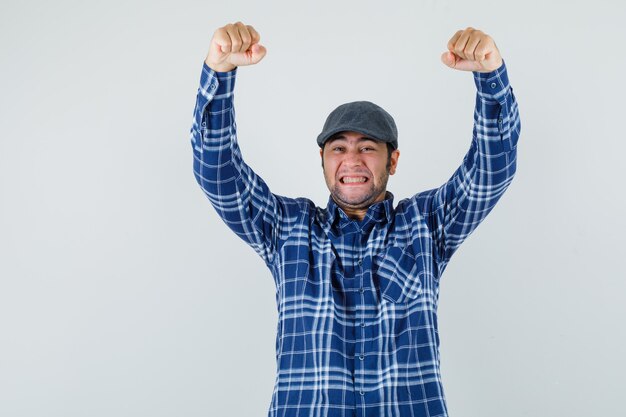 Jonge man die winnaargebaar in overhemd, pet toont en zalig, vooraanzicht kijkt.