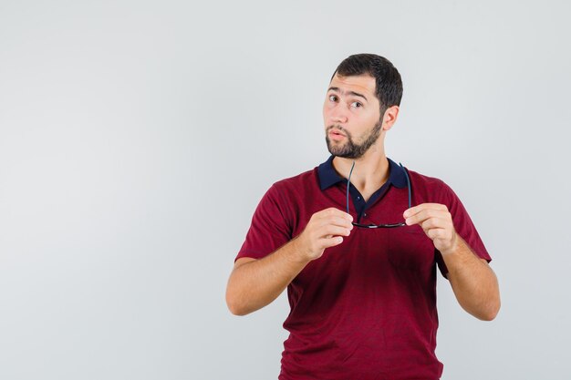 Jonge man die zijn bril in rood t-shirt houdt en nadenkend, vooraanzicht kijkt.