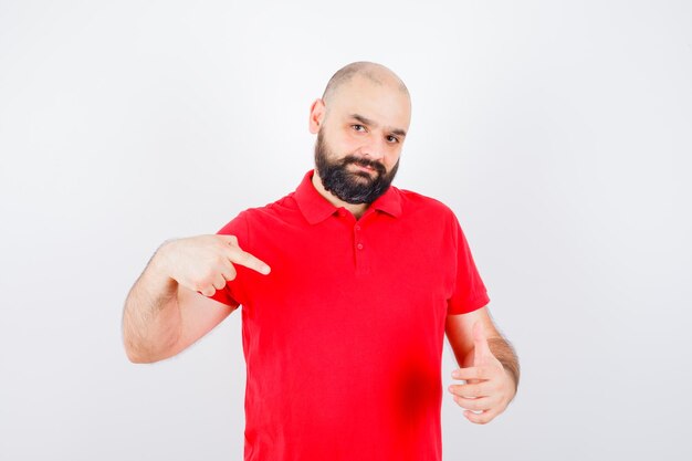 Jonge man die zijn duim opsteekt terwijl hij ernaar wijst in een rood shirt en er zelfverzekerd uitziet. vooraanzicht.