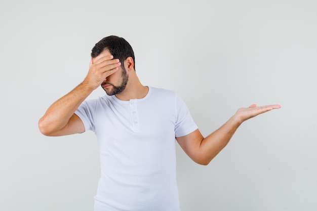 Jonge man die zijn gezicht bedekt met zijn hand terwijl hij opzij wijst in een t-shirt en er ontevreden uitziet, vooraanzicht.