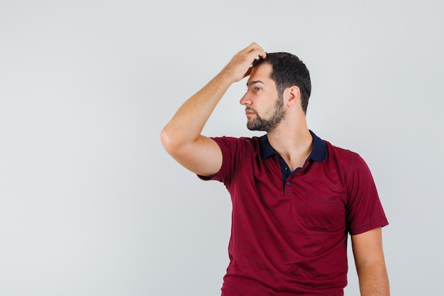 Jonge man die zijn hoofd krabt terwijl hij opzij kijkt in een rood t-shirt en verward kijkt. vooraanzicht.