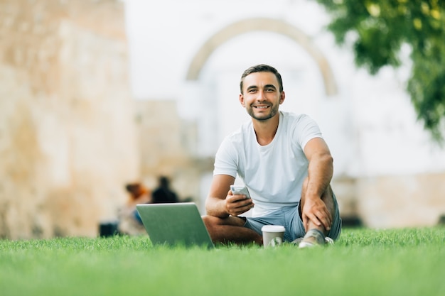 Jonge man die zijn laptop met koffie gebruikt om op het gras te gaan werken