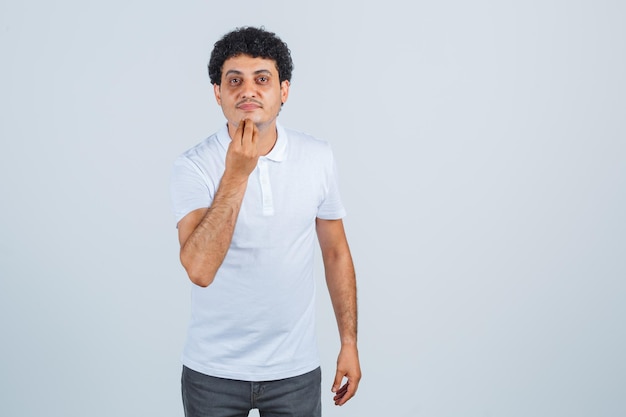 Jonge man doet Italiaans gebaar in wit t-shirt, broek en ziet er zelfverzekerd uit. vooraanzicht.