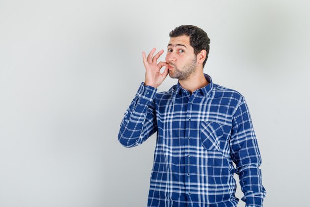 Jonge man doet slotgebaar op lippen in ingecheckte overhemd en kijkt serieus