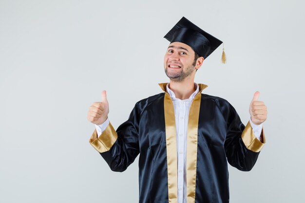 Jonge man dubbele duimen opdagen in afgestudeerde uniform en op zoek zelfverzekerd, vooraanzicht.