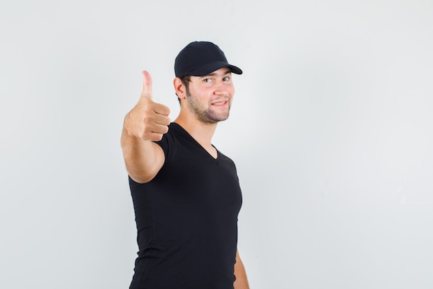 Jonge man duim opdagen in zwart t-shirt, pet en op zoek vrolijk.