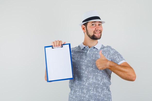 Jonge man duim opdagen terwijl hij zijn notitieboekje in t-shirt, hoed vasthoudt en zelfverzekerd kijkt, vooraanzicht.