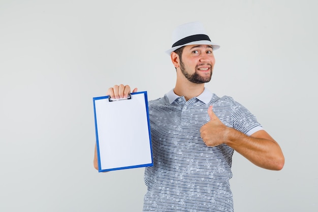 Jonge man duim opdagen terwijl hij zijn notitieboekje in t-shirt, hoed vasthoudt en zelfverzekerd kijkt, vooraanzicht.