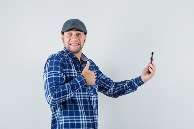 Jonge man duim opdagen tijdens het maken van videogesprek in shirt, pet en op zoek blij, vooraanzicht.