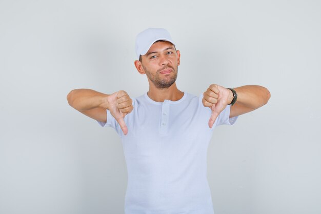 Jonge man duimen omlaag in wit t-shirt, pet en op zoek naar ontevreden, vooraanzicht.