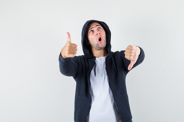 Jonge man duimen op en neer in t-shirt, jasje en op zoek naar besluiteloos, vooraanzicht.