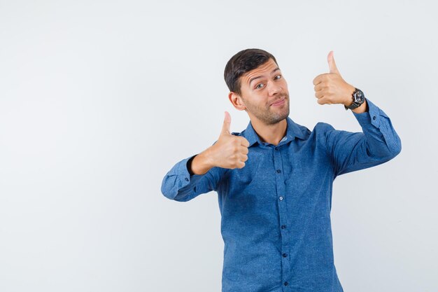 Jonge man duimen opdagen in blauw shirt en op zoek gelukkig. vooraanzicht.