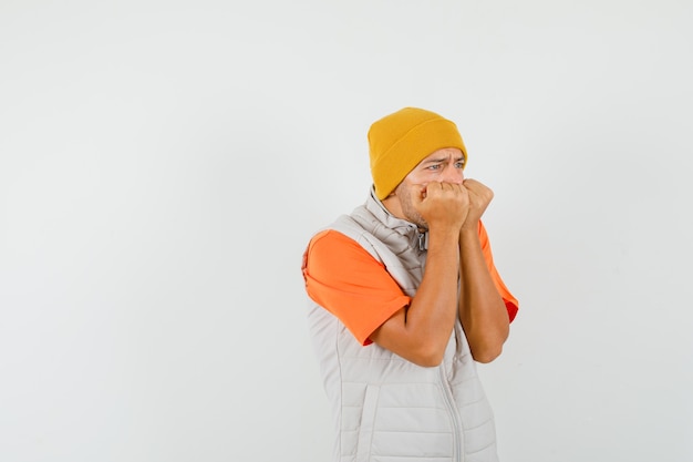 Jonge man emotioneel vuisten bijten in t-shirt, jas, hoed en op zoek bang. vooraanzicht.