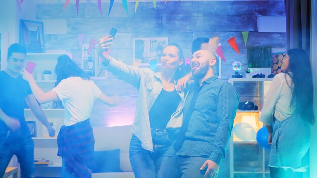 Jonge man en vrouw die grappige gezichten trekken terwijl ze selfies nemen op een feestje met neonlichten en rook.