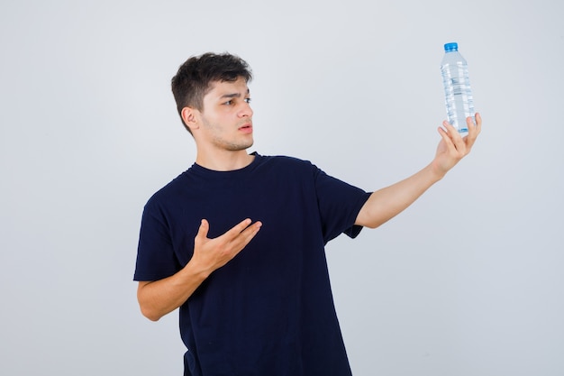 Jonge man fles water in zwart t-shirt tonen en verbaasd op zoek. vooraanzicht.