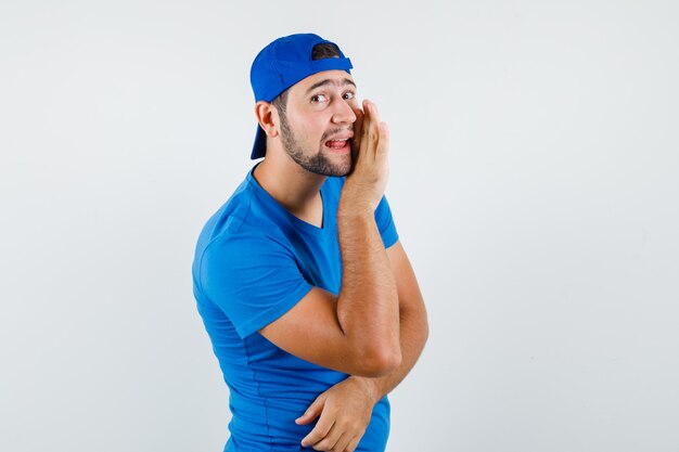 Jonge man geheim achter hand in blauw t-shirt en pet en op zoek roddels