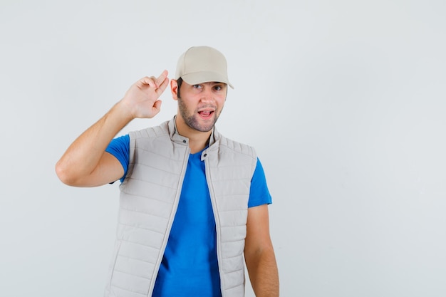 Jonge man groet gebaar met vingers in t-shirt, jasje, pet en op zoek zelfverzekerd. vooraanzicht.