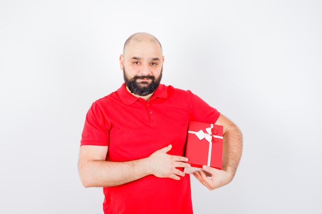 Jonge man houdt cadeau in rood shirt en ziet er tevreden uit. vooraanzicht.