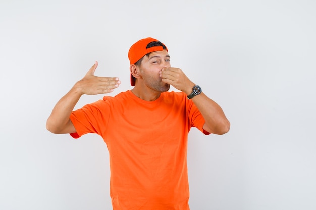 Jonge man houdt de adem in met vingers op de neus in oranje t-shirt en pet en kijkt walgend