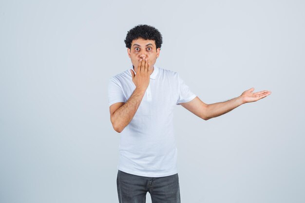 Jonge man houdt de hand op de mond terwijl hij iets in een wit t-shirt, broek laat zien en verrast kijkt, vooraanzicht.