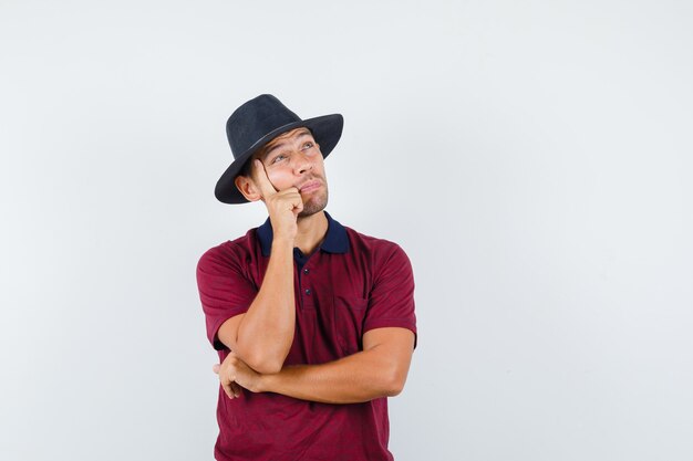 Jonge man houdt de hand op zijn kin terwijl hij wegkijkt in een rood shirt, zwarte hoed en verbaasd kijkt. vooraanzicht.