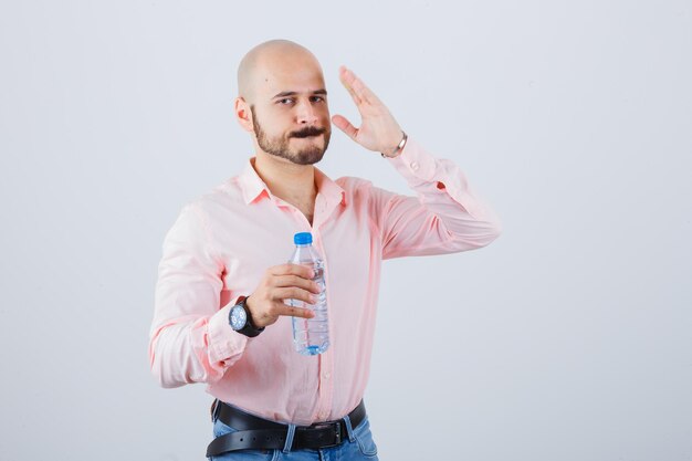 Jonge man houdt hand in de buurt van gezicht in shirt, spijkerbroek en ziet er serieus uit, vooraanzicht.