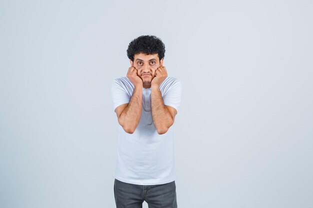 Jonge man houdt handen op wangen in wit t-shirt, broek en kijkt angstig. vooraanzicht.