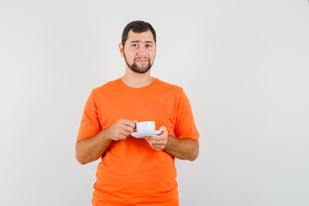 Jonge man houdt kop met schotel in oranje t-shirt en ziet er zachtaardig uit. vooraanzicht.